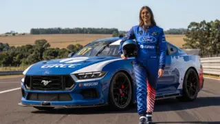 Rafaela Ferreira, piloto brasileira, posa ao lado de um carro de corrida Ford Mustang #7 da equipe Cavaleiro Sports, com o capacete em mãos.